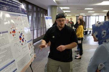 A student makes a presentation beside a poster at the 2024 Student Conference for Research, Professional Activities, and Creative Arts