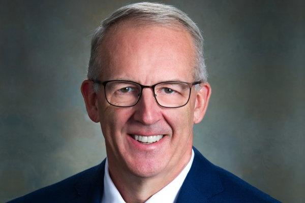 Greg Sankey, a man with short cut gray hair in glasses and blue suit, smiles at camera.